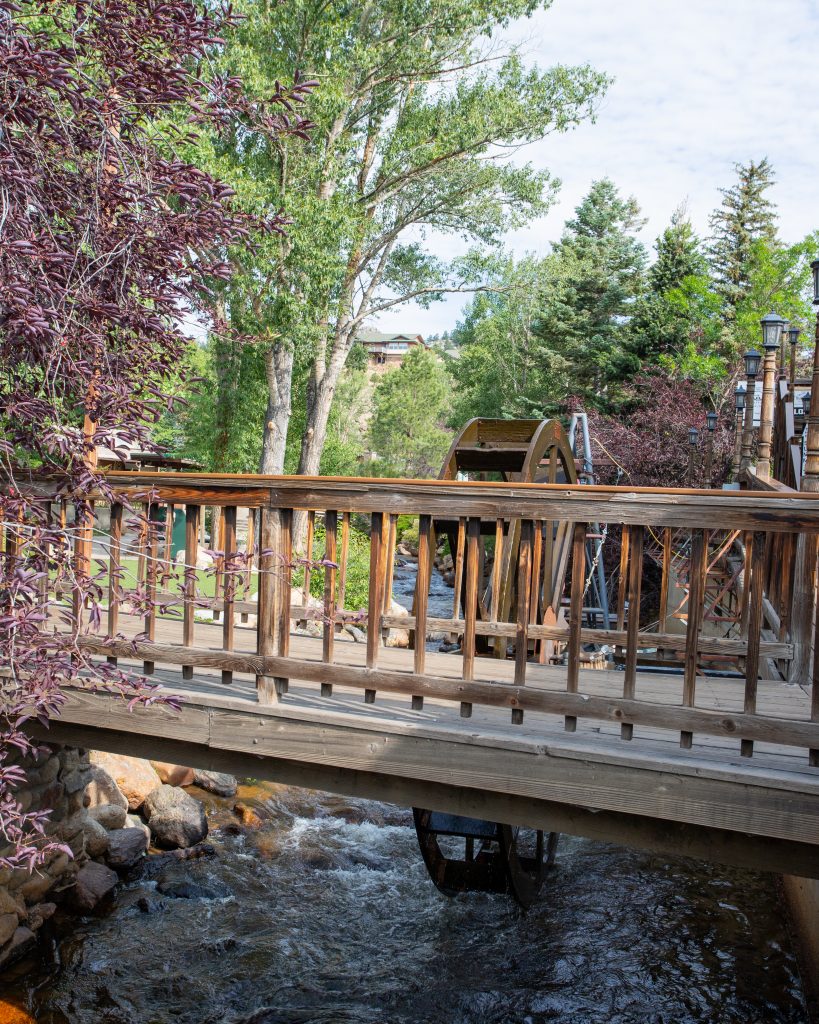 A water wheel sits behind a bridge under an archway of trees in Downtown Estes Park